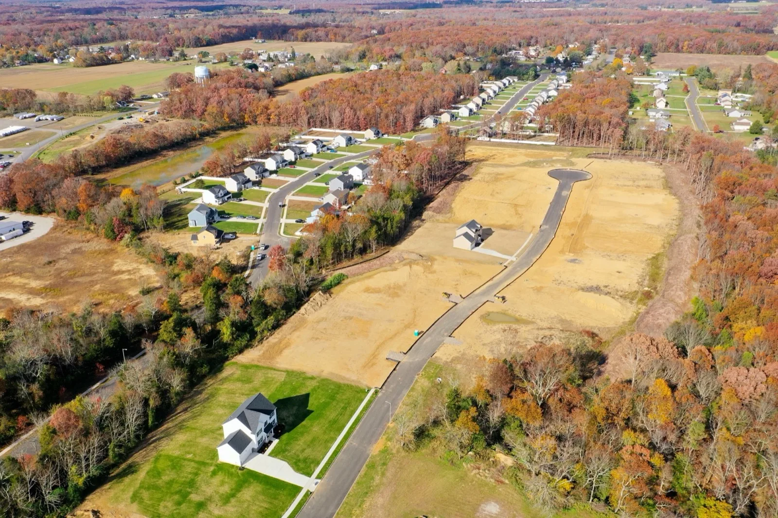 An aerial view of a new street cut through graded homesites, with new houses along it and autumn woods around